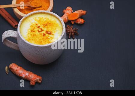 Detox Gelbwurz latte, goldene Milch trinken in einem Becher, Pulver und Wurzeln auf schwarzem Hintergrund, Nahaufnahme Stockfoto