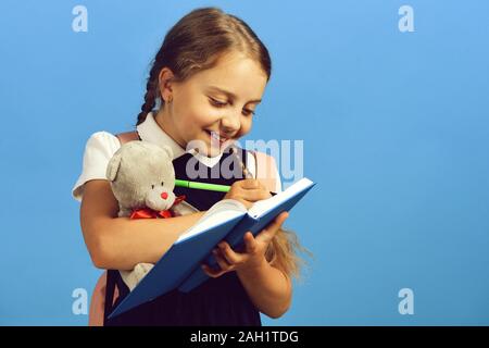 Mädchen hält Teddybären und schreibt in Blau notebook. Zurück zu Schule und Bildung Konzept. Schule Mädchen mit glücklichen Gesicht auf blauem Hintergrund isoliert. Schüler in der Schule Uniform mit Zopf und rosa Rucksack Stockfoto