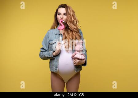 Lustige casual schwangere Mädchen mit Hase Spielzeug süsse bonbons am gelben studio Hintergrund posiert Stockfoto