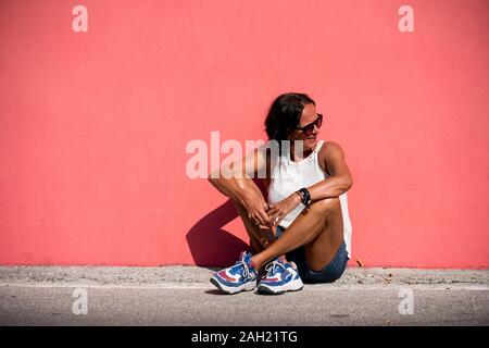 Attraktive ältere Frau mit Sonnencreme mit Sonnenbrille gegen ein helles Licht rosa monochromen Wand lehnt. Sunny sunshine Tag einer Person älter reife weibliche. Stockfoto