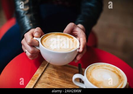 In der Nähe der Hände Person in Lederjacke mit einer Tasse heißen Cappuccino und Latte Art im Cafe. Person Kaffee trinken mit schönen Schaum. Cup o Stockfoto