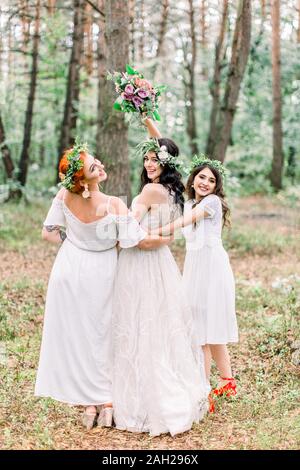 Braut und Brautjungfern in weißen Kleidern auf dem Wald Hintergrund im Freien posieren. Braut ihren Blumenstrauß Holding. Rustikale Hochzeit im Wald. Stockfoto