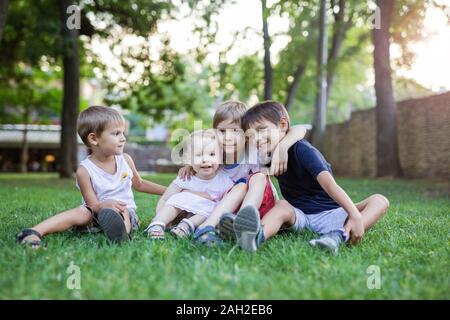Drei Jungen und Kleinkind Mädchen im Sommer Park. Freunde oder Geschwister sitzen auf grünem Gras. Stockfoto