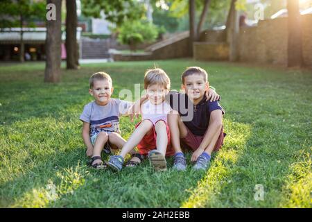 Drei glückliche Jungs im Sommer Park. Freunde oder Geschwister sitzen auf grünem Gras. Stockfoto