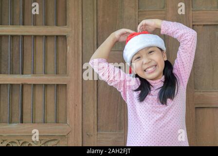 Süße asiatische Mädchen mit weihnachtsmütze Lächeln auf altem Holz wand hintergrund, Weihnachten Konzept Stockfoto