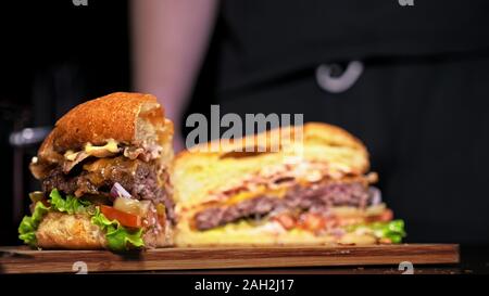 Schneiden Handwerk Burger ist Kochen auf schwarzem Hintergrund. Bestehen: Sauce, Salat, Tomaten, Roten Zwiebeln, Gurke, Käse, Speck, Brötchen und marmoriertes Fleisch Rindfleisch. Stockfoto