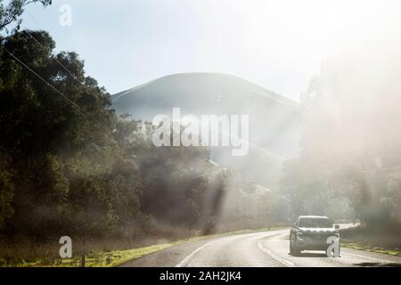 Blick durch die Windschutzscheibe der geschwungenen Asphalt-Landstraße, mit dem Auto, das unter grünen Wald mit Bergen im Hintergrund am sonnigen Tag führt Stockfoto