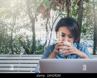 Schöne happy asiatische Frau kurze Haare trägt blaue Jeans shirt Trinken weiße Tasse heißen Kaffee und Suchen auf Laptop im Cafe mit grünen Bac Stockfoto