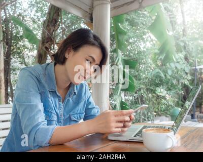 Schöne happy asiatische Frau mit Handy mit Laptop und eine Tasse Kaffee auf hölzernen Tisch in Coffee Shop. Stockfoto