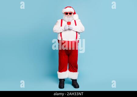 Volle Länge Foto von modernen Stil lustig funky fat Santa Claus mit großen Bauch nutzen Handy Suche Winter Sales weißen Overalls Rot tragen Stockfoto