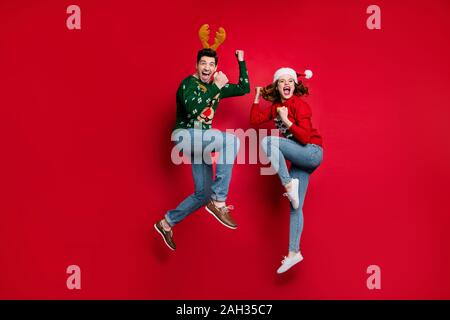 Voller Körper Foto von überrascht springen Paar von x-mas Preise tragen hässliche ornament Jumper und headwear isolierten roten Farbe Hintergrund begeistert Stockfoto