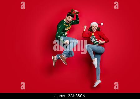 Voller Körper Foto von überrascht springen Paar von x-mas Preise tragen hässliche ornament Jumper und headwear isolierten roten Farbe Hintergrund begeistert Stockfoto