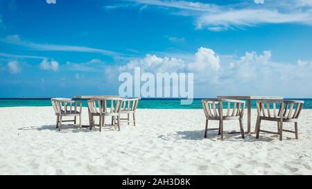 Stühle und Tische auf den weißen Strand der Malediven mit dem türkisblauen Meer im Hintergrund und Wolken am blauen Himmel Stockfoto