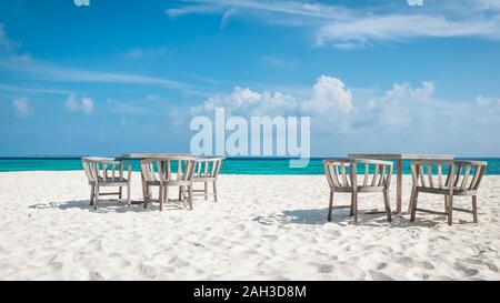 Stühle und Tische auf den weißen Strand der Malediven mit dem türkisblauen Meer im Hintergrund und Wolken am blauen Himmel Stockfoto