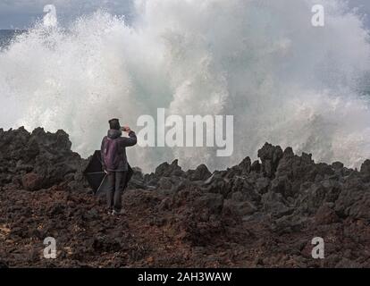 Eine einsame weibliche nimmt ein Foto mit einem Handy eine große Welle krachend gegen die Felsen. Sie scheint nicht die gefährliche Position, die Sie in ist. Stockfoto