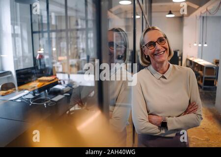 Portrait von lächelnden reife Geschäftsfrau lehnte sich gegen Glasscheibe im Büro Stockfoto