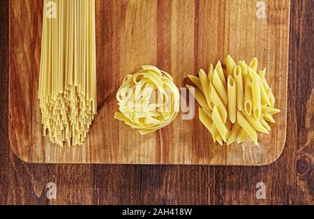 Verschiedene Arten von Pasta auf Holztisch mit Schneidebrett, Draufsicht. Pappardelle, Spaghetti, Penne Pasta auf dunklem Holztisch. Stockfoto