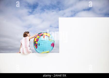 Kleines Mädchen sitzt an der Wand, mit Arm um aufblasbarer Globus, Rückansicht Stockfoto