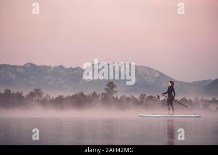 Frau Stand up Paddeln auf dem See Kirchsee im Morgennebel, Bad Tölz, Bayern, Deutschland Stockfoto