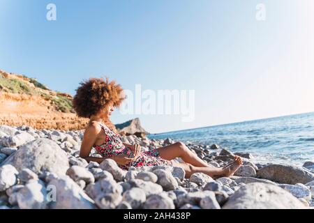 Junge Frau Sommer Kleid tragen und sitzen auf steinigem Strand Stockfoto