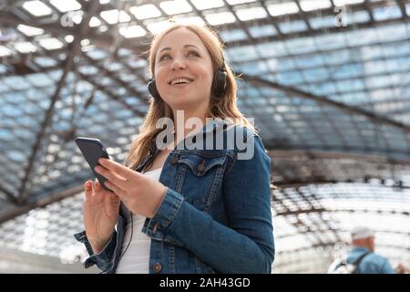 Lächelnde Frau mit Smartphone und Kopfhörer am Bahnhof, Berlin, Deutschland Stockfoto