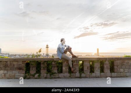 Mann sitzt auf einer Wand auf Ausblick über der Stadt mit Blick auf den Hafen, Barcelona, Spanien Stockfoto