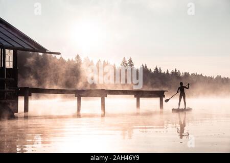 Silhouette einer Frau Stand up Paddeln auf dem See Kirchsee im Morgennebel, Bad Tölz, Bayern, Deutschland Stockfoto