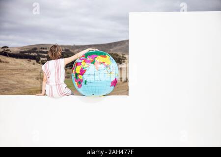 Kleines Mädchen sitzt an der Wand, mit Arm um aufblasbarer Globus, Rückansicht Stockfoto