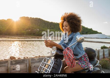 Junge Frau sitzt auf einem Roller- und Sie ihr Smartphone bei Sonnenuntergang, Ibiza Stockfoto