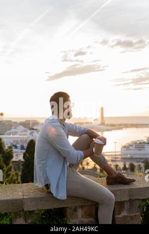 Mann sitzt auf einer Wand auf Ausblick über der Stadt mit Blick auf den Hafen, Barcelona, Spanien Stockfoto