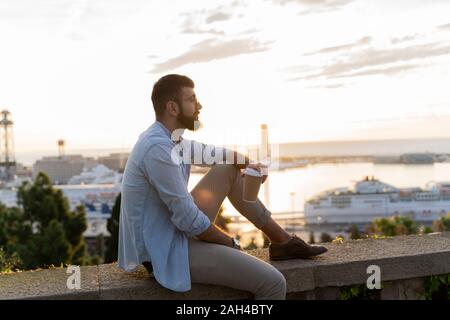 Mann sitzt auf einer Wand auf Ausblick über der Stadt mit Blick auf den Hafen, Barcelona, Spanien Stockfoto