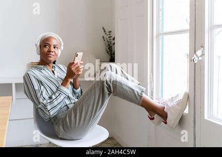 Lächelnde junge Frau mit Kopfhörer und Handy am Fenster Stockfoto