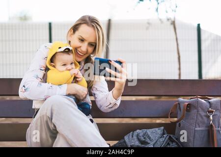 Glückliche Mutter mit Baby auf einer Parkbank ein selfie Stockfoto
