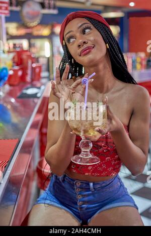 Junge Frau mit geflochtenem Frisur sitzt auf einem Bar, trinken Sie einen Cocktail mit Strohhalm Stockfoto