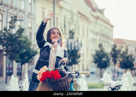 Schöne junge lächelnde Frau mit Fahrrad und winkende Hand auf Straße der Stadt. Schönheit, Geste und Lifestyle Konzept Stockfoto