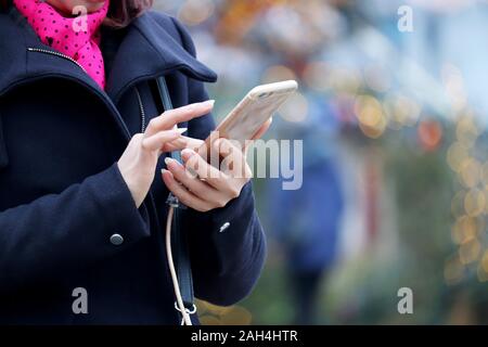 Frau in einem Winter Fell steht mit Smartphone in den Händen auf Weihnachtsbeleuchtung Hintergrund auf einer winter Straße. Neues Jahr Feier in einer Stadt Stockfoto