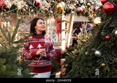 Glückliche Frau in einem roten Pullover mit Lollipop genießt die Magie der Weihnachtszeit stehen auf einer Straße in neuen Jahr Baum Hintergrund Stockfoto