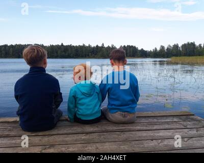 Drei Jungen verschiedenen Alters in blauer Pullover in verschiedenen Farben sitzen auf einem warmen Ponton aus Holz, Beine baumeln, ihren Rücken auf den Betrachter, auf den See und die grünen Wald dahinter suchen. Stockfoto