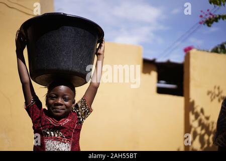 Kleine Afrikanische Mädchen, die zu Hause einen großen Wassereimer auf dem Kopf Stockfoto