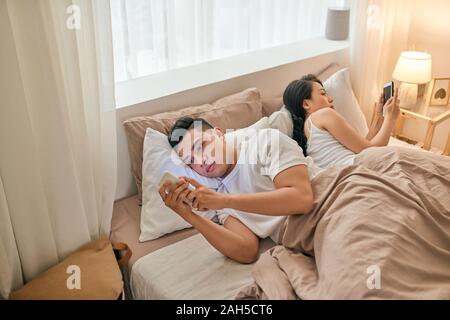 Junges Paar im Bett mit Phone lügen Rücken zueinander. Stockfoto