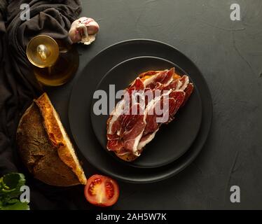Brot mit Schinken auf schwarzem Teller und rustikalen Hintergrund getoastet, im Hintergrund Artisan Brot, frische Tomaten und Olivenöl Stockfoto
