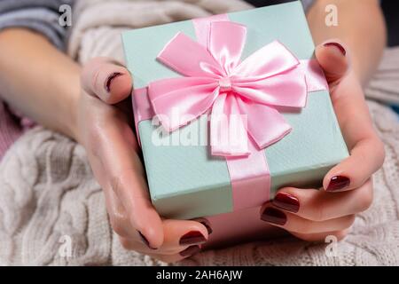 Mädchen Hände halten einen blauen Geschenkbox mit rosa Krawatte Schleife auf wollpullover Hintergrund. Weibliche Hände mit braunen Nägel Polnisch auf die Finger Stockfoto