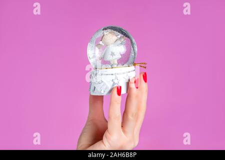 Frau Hand ornament Kugel mit Schnee auf rosa Hintergrund im Studio isoliert. Weibliche Hände mit roten Nägeln Polnisch auf die Finger. Weihnachten Konzept Stockfoto