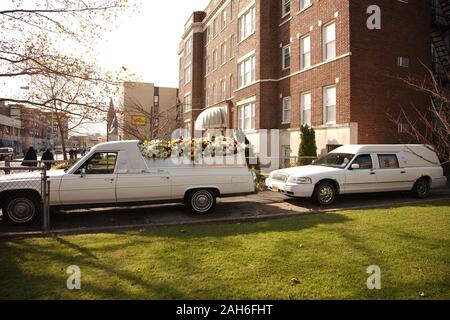 Ein weißer Trauerblumenwagen, der vor einem passenden weißen Leichenwagen geparkt ist, Seitenansicht beider Fahrzeuge, bereit für eine Trauerprozession. Stockfoto