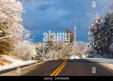 Eine zweispurige Landstraße schlängelt sich durch das Land im Winter. Mit gelben Linien in seiner Mitte, gesäumt von Stromleitungen, Bäumen und einem kleinen Haus. Stockfoto