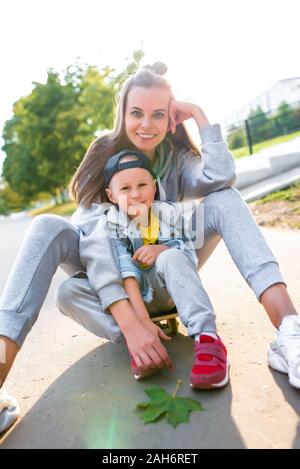 Frau Mutter sitzt auf Skateboard, lustige Leute freuen und lachen, kleine Junge 3-5 Jahre, im Sommer in der Stadt, im Herbst, Gefühle der Liebe, Pflege Stockfoto