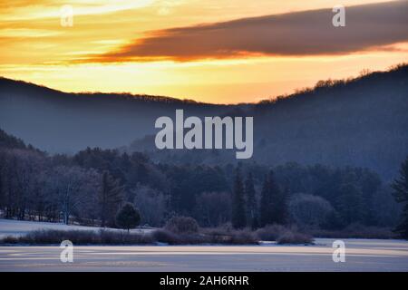 Kalte Sonnenaufgang über Baum bedeckte Hügel und gefrorenes Wasser mit Schnee am Roten Haus See, Allegany State Park, New York Stockfoto