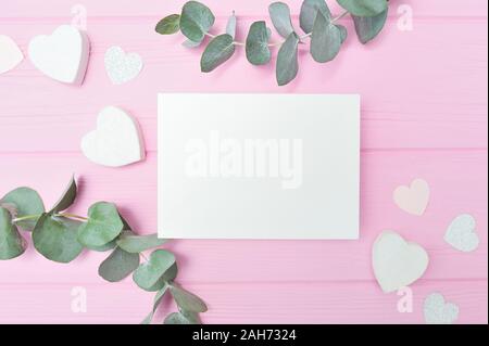 Valentines Tag oder Hochzeit mockup Szene mit leere Karte, Eukalyptus Blätter frame Papier Herzen Konfetti- und Holz- rosa Hintergrund, leeren Raum für Ihre Stockfoto