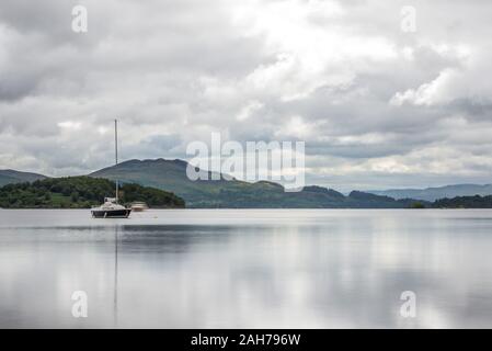 Ein schottischer See umgeben von Bäumen und Vegetation mit einem Segelboot in der Mitte und fernen Bergen im Hintergrund, unter einem bewölkten Himmel Stockfoto