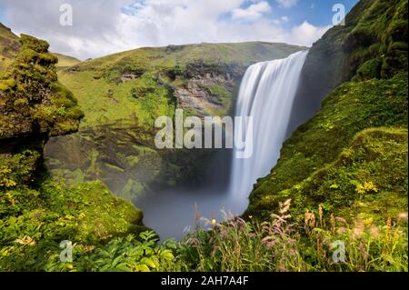 Lange Exposition des isländischen Wasserfalls von Skogafoss, umgeben von Klippen mit grünem Moschus, unter einem blauen Himmel mit Wolken bedeckt Stockfoto